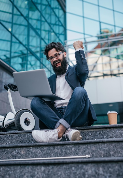 Bebaarde volwassen zakenman met laptop buiten het vieren van succes