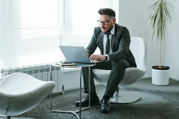 Bebaarde zakenman in elegant pak zittend in een stoel voor laptop op kantoor en het verzenden van e-mail naar collega of zakenpartner