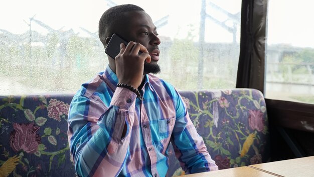 Bebaarde zwarte jonge man praat op mobiele telefoon. Afro-Amerikaanse man belt op smartphone die in café zit te wachten op zijn bestelling. Kerel in zomertentcafé zittend op de bank.