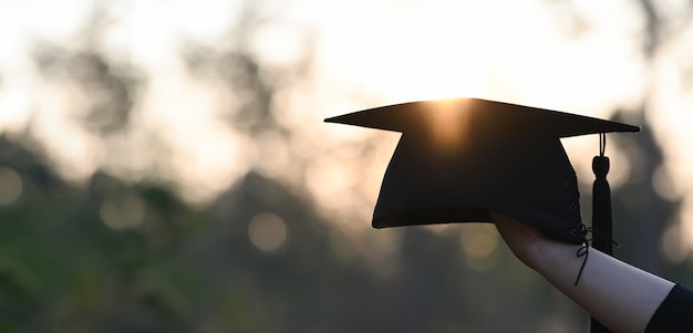 Bebouwd beeld van universitaire studentenhand die een graduatiehoed in openlucht overhandigen met zonsondergang als achtergrond.