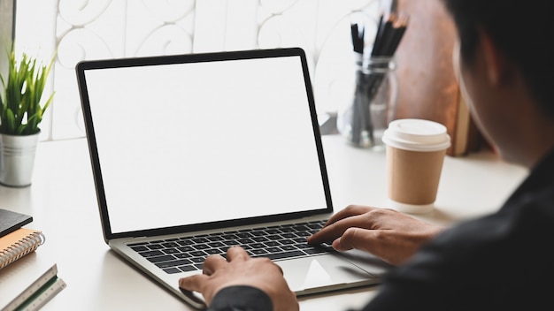 Bebouwd beeld van zakenmanhanden die op computerlaptop typen met het witte lege scherm dat het zetten van ordelijk bureau dat omringd met boeken, potloodhouder, koffiekop en potplant.