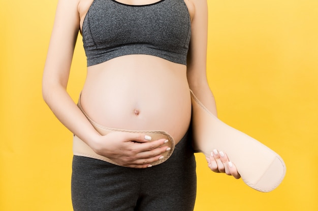 Bebouwd beeld van zwangere vrouw op het derde trimester die op zwangerschapsriem zetten bij gele achtergrond. Orthopedisch buikondersteuningsriemconcept met exemplaarruimte