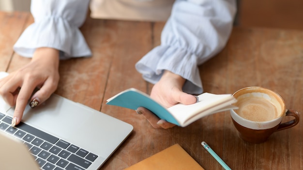 Bebouwd schot van een meisje die op laptop typen terwijl het kijken op haar notitieboekje op houten bureau