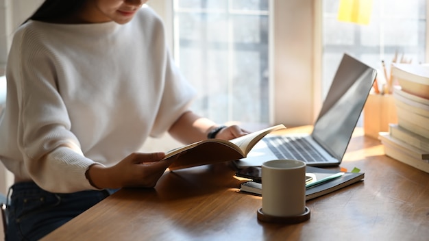 Bebouwd schot van jonge ontwerpervrouw die een boek lezen terwijl het zitten aan het moderne bureau met zonlicht door het venster als Ontspannende tijd na beëindigd werk. Pauze.