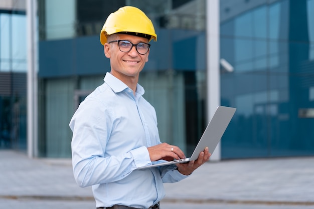 Bedrijf, architect met behulp van laptop geklede helm brillen buitenshuis kantoorgebouw achtergrond blanke mannelijke ingenieur controle bouwproces buiten