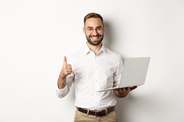 Bedrijf. Jonge succesvolle mannelijke ondernemer die duim-omhoog toont tijdens het werken aan laptop, die zich over witte achtergrond bevindt.