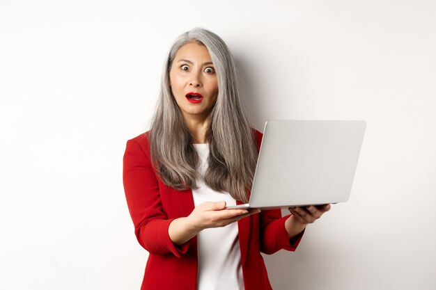 Bedrijf. Onder de indruk Aziatische oude vrouw die op laptop werkt, camera verbaasd staren, die zich over witte achtergrond bevindt.