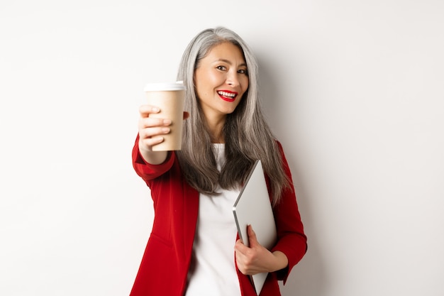 Bedrijf. Vrolijke Aziatische vrouwenmanager die u kop van koffie geeft en glimlacht, die zich met laptop in hand, witte achtergrond bevindt.