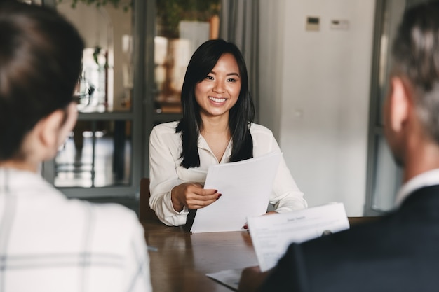 Bedrijfs-, carrière- en rekruteringsconcept - jonge Aziatische vrouw die glimlacht en cv vasthoudt, tijdens het interviewen als kandidaat voor een baan in een groot bedrijf