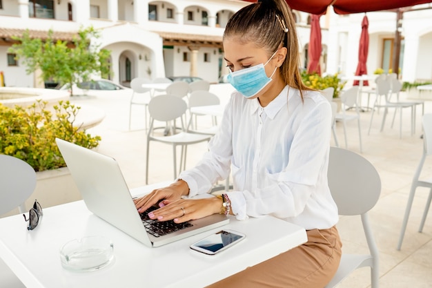 Bedrijfs jonge vrouw buiten met laptop op bartafel die beschermend masker tegen coronavirus pandemie draagt. nieuwe normale freelance job activiteit zelfstandig ondernemer overal werkzaam met mobiel internet