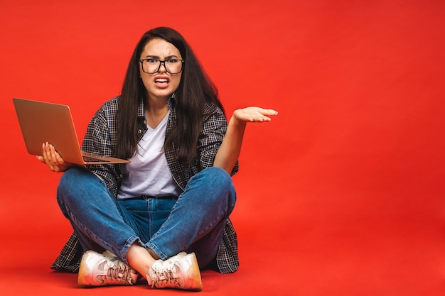 Bedrijfsconcept portret van boze vermoeide brunette vrouw in casual zittend op de vloer in lotus houding en met laptop geïsoleerd op rode achtergrond