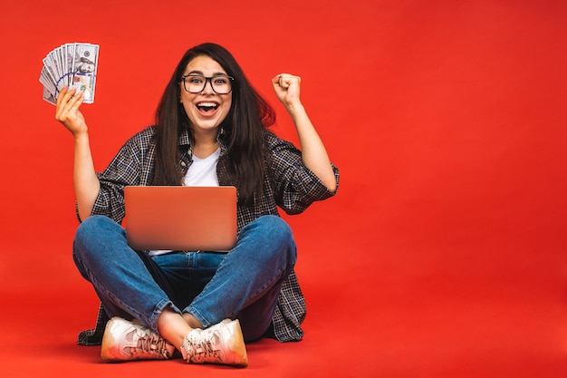 Bedrijfsconcept portret van gelukkige brunette vrouw in casual zittend op de vloer in lotus houding en met laptop geïsoleerd op rode achtergrond Holding geld rekeningen