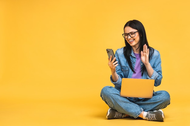 Bedrijfsconcept portret van gelukkige jonge vrouw in casual zittend op de vloer in lotus houding en met laptop geïsoleerd op gele achtergrond met behulp van mobiele telefoon