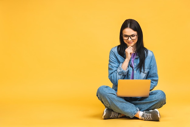 Bedrijfsconcept portret van gelukkige jonge vrouw in casual zittend op de vloer in lotus houding en met laptop geïsoleerd op gele achtergrond