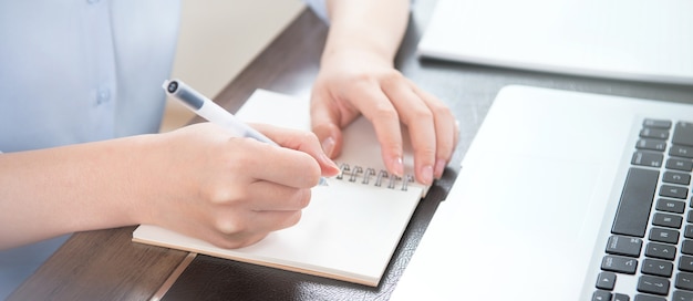 Bedrijfsconcept. Vrouw neemt nota voor project met laptop en rapporteert in bureau. Achtergrondverlichting, verblindend effect, close-up, zijaanzicht, kopieerruimte