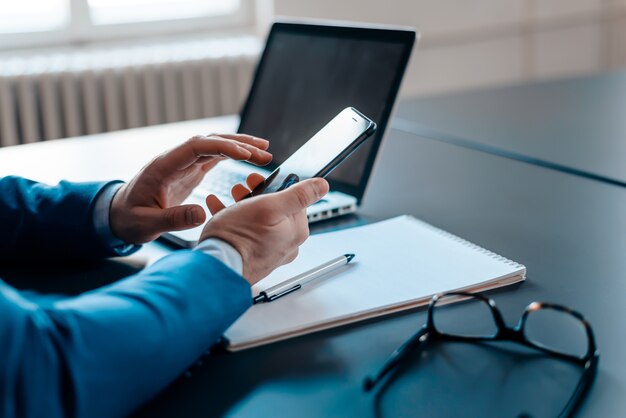 Bedrijfsmens die smartphonezitting bij bureau met laptop, pen, notitieboekje en oogglazen gebruiken.