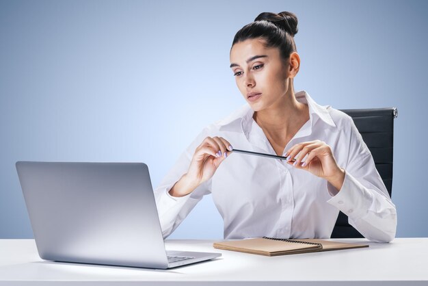 Bedrijfsproces en baanconcept met mooie jonge vrouw die aan een wit bureau zit en naar het scherm van de laptop kijkt en een potlood in handen draagt op een abstracte lichtblauwe muurachtergrond