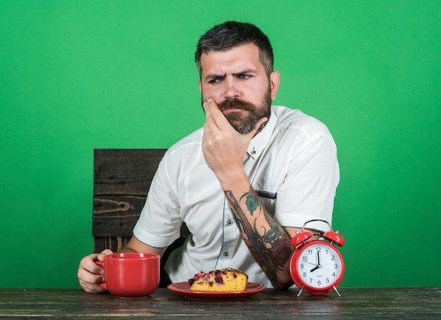 Bedroefde bebaarde man die ontbijt eet, kopje cappuccino of koffie heeft en een stuk smakelijke taart op bord. Mensen, vrije tijd en voedsel concept - knappe man in wit overhemd zit aan bureau ontbijt te eten.