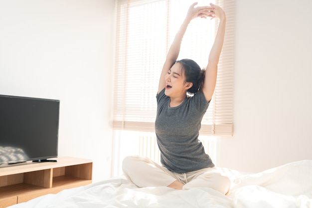 Bedroom concept a pretty girl looking sleepy yawning while stretching her hand on the white comfortable bed.