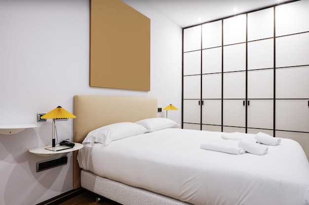 Bedroom with a double bed with a white feather duvet with white shelves as bedside tables with twin lamps and a large builtin wardrobe with white wooden doors with bars