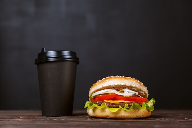 写真 黒い紙の上のコーヒーとベーコンのビーフバーガー木製のテーブルの上のカップ。カフェメニューデザインコンセプト