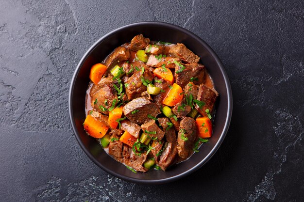 Photo beef meat and vegetables stew in black bowl slate background top view