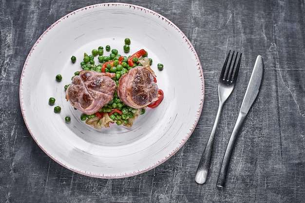 Foto medaglioni di manzo avvolti in pancetta, servire con piselli e su un piatto su fondo di legno.