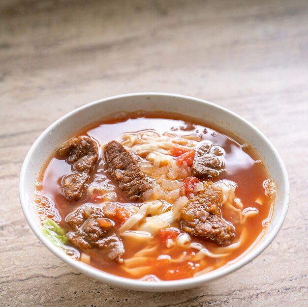 Beef noodle ramen meal with tomato sauce broth in bowl on\
bright wooden table famous chinese style food in taiwan close up\
top view copy space