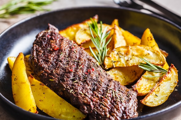 Beef steak and baked potato wedges with rosemary on black plate, dark background.