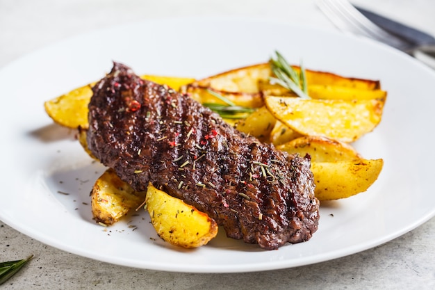 Foto bistecca di manzo e spicchi di patate al forno con rosmarino in zolla bianca.