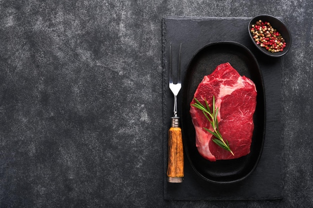 Beef steak fresh raw beef rib eye steak with fork rosemary salt\
and pepper on a grill pan on dark grey or black stone background\
shop advertising idea top view mockup for design idea