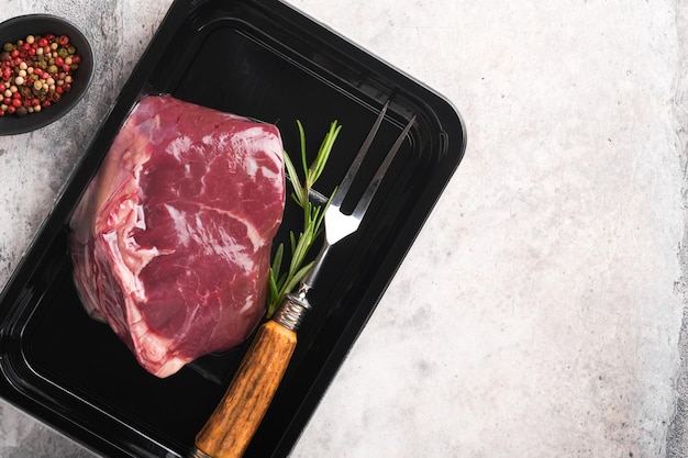 Beef steak fresh raw beef steak with fork and rosemary in
vacuum black plastic pack on grey stone background vacoom package
top view mockup for design idea