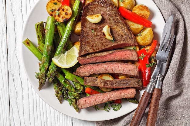 beef steak with barbequed new baby potatoes, asparagus, lemon and red pepper on a white plate with barbecue sauce on a wooden table, flat lay, macro