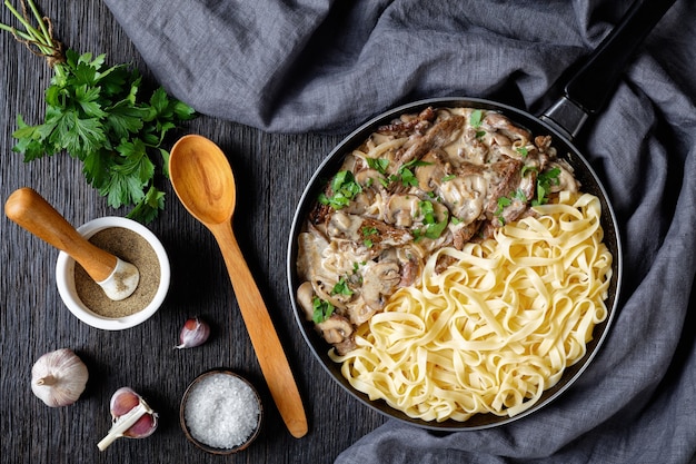 Beef stroganoff cooked with sour cream mushrooms sauce, served with egg noodles in a skillet on a dark wooden table, flat lay, horizontal view from above