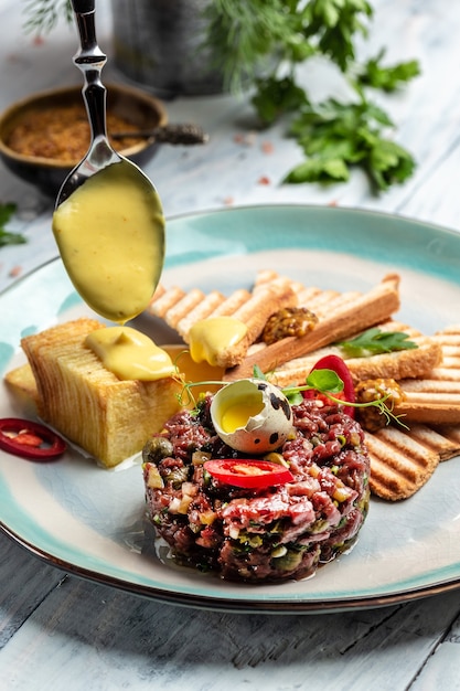 Beef tartar with egg. Tartar with raw quail egg yolk, capers, pickled cucumber with bread toasts served on plate. Raw tartare meat. French cuisine