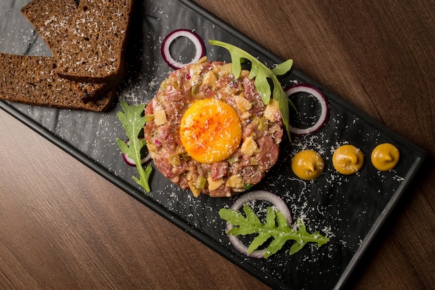 Beef tartare with greens onions egg mustard and bread on a black plate Tartare top view