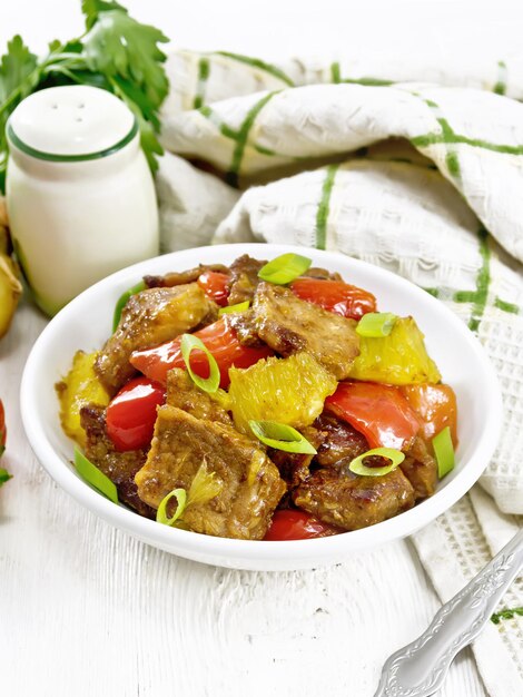 Beef with oranges, bell peppers and ginger root in bowl, a napkin and a fork on wooden board background