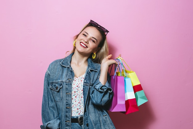 Beeld dat van een mooi blondemeisje met zonnebril glimlacht, het winkelen zakken over roze muur houdt