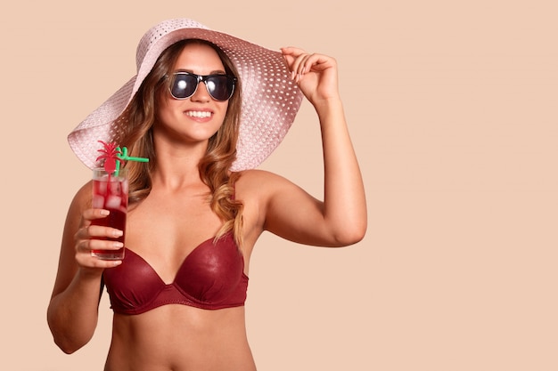 Beeld die van vrolijke knappe toerist strohoed, rood zwemmend kostuum en zonnebril dragen, opzij kijkend, genietend van de zomervakantie, houdend glas van koude cocktail. Ruimte voor reclame kopiëren.