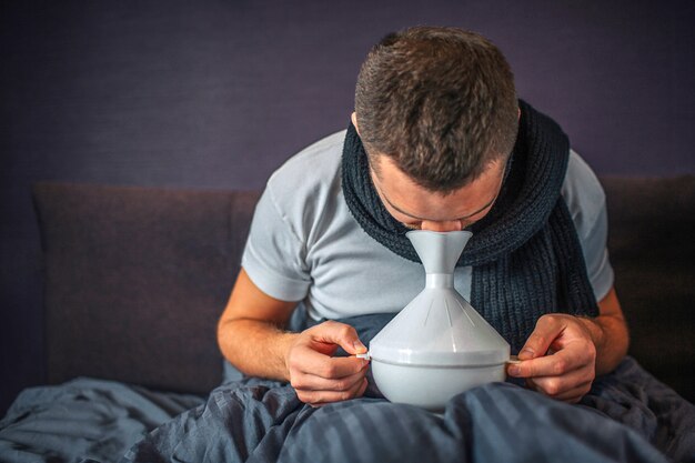 Beeld van de mens die inhalator gebruikt voor behandeling en om beter te worden. Hij is ziek en ziek. Guy houdt het witte ding voorzichtig en met beide handen vast. Hij zit op bed.