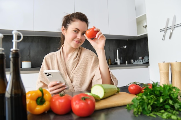 Beeld van een jonge, mooie vrouw met tomaat die in de keuken zit met een smartphone-snijplank en