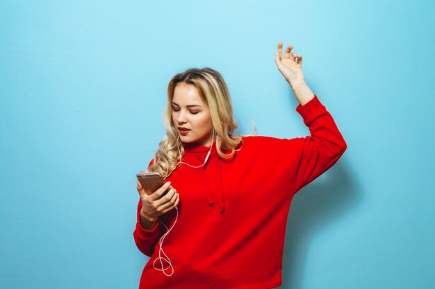 Beeld van een mooi blond opgewekt meisje dat aan muziek in hoofdtelefoons luistert en over blauwe muur danst