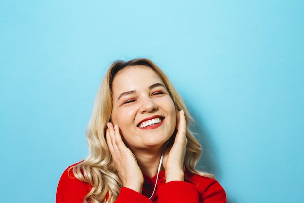 Beeld van een mooi blond opgewekt meisje dat aan muziek in hoofdtelefoons luistert en over blauwe muur danst