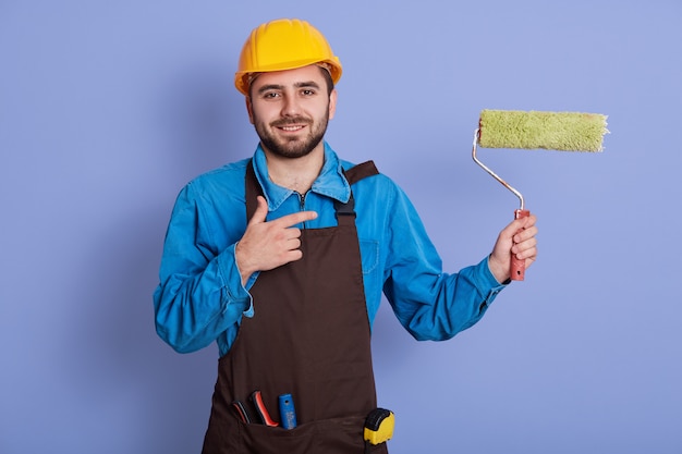 Beeld van energieke charismatische knappe jonge handarbeider die oprecht glimlacht, aan het werk is, gebaar maakt, rol in één hand houdt, richting met wijsvinger toont. beroep concept.