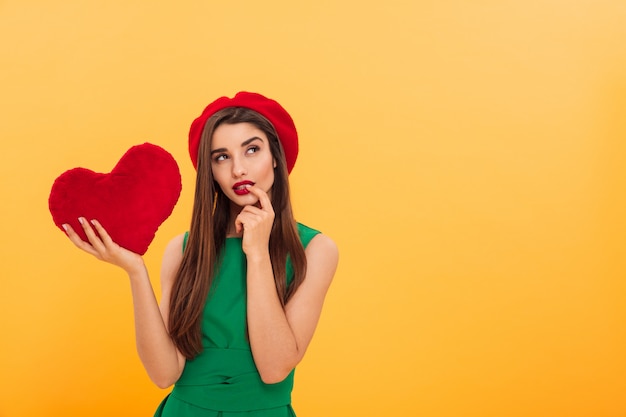 Beeld van nadenkende jonge vrouw status geïsoleerd over het gele hart van de muurholding. Opzij kijken.