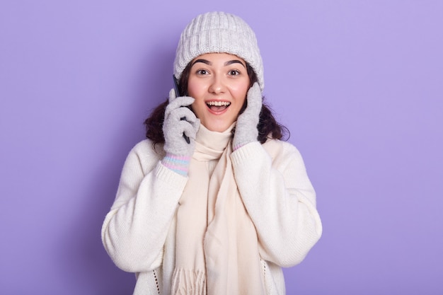 Beeld van verraste emotionele jonge dame die met donker krullend haar direct camera bekijken, smartphone dichtbij oor houden, opheffend haar handen