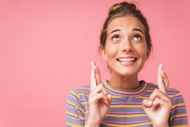 Beeldclose-up van glimlachende Kaukasische vrouw die gestreept t-shirt draagt dat omhoog kijkt met gekruiste vingers geïsoleerd over roze muur