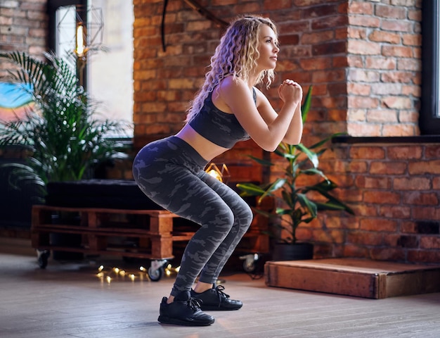 Beentrainingen voor vrouwen. Blonde sportieve vrouw doet squats in een kamer met loft interieur.