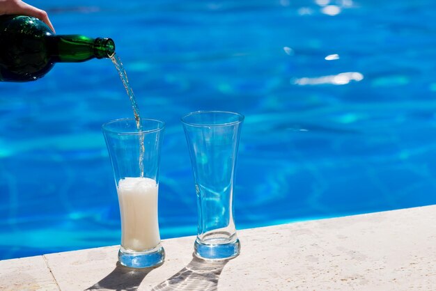 Beer is pouring into a two glasses from green bottle near the pool