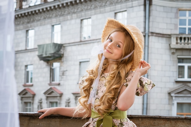 Beetje mooi meisje in jurk en hoed poseren op het balkon in de stad